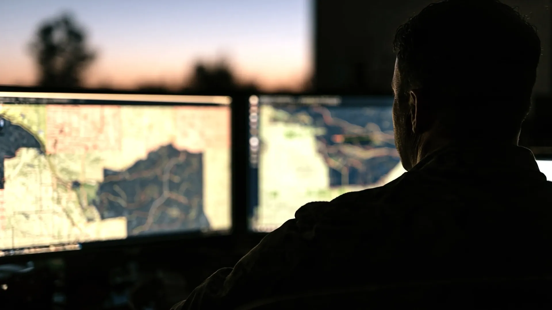An analyst at two map screens at dusk, seen from behind.